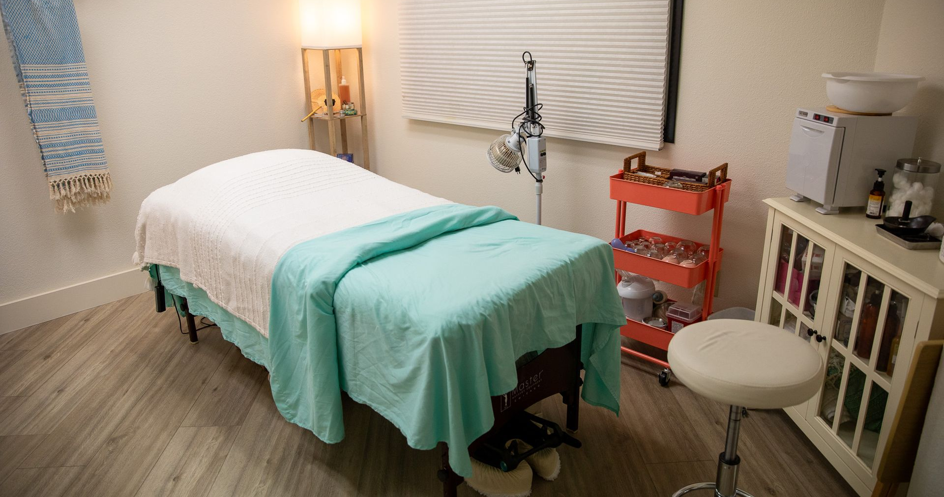 Massage therapy room with bed, turquoise and white linens, lamp, and stool.