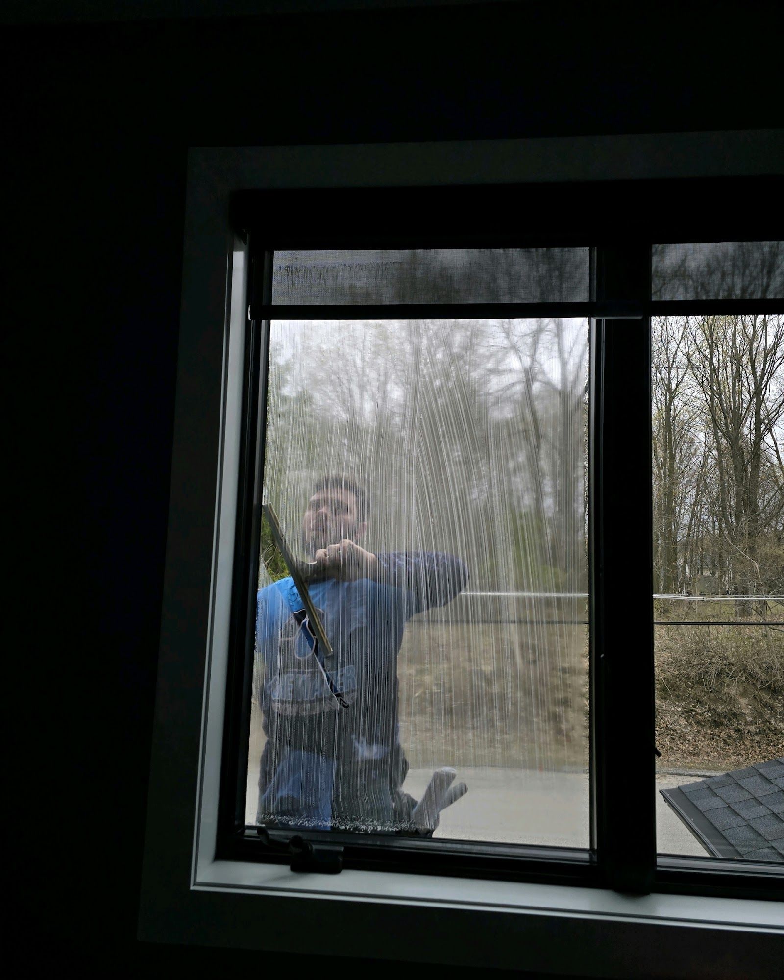 Person cleaning a window with a squeegee. They're visible through the window, in a blue shirt, with a gray backdrop.