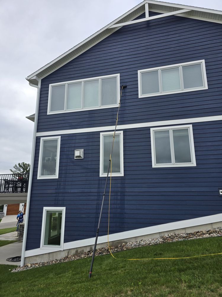 A person washing windows on a blue house using a long pole with a brush. The house is two stories and sits on a grassy hill.