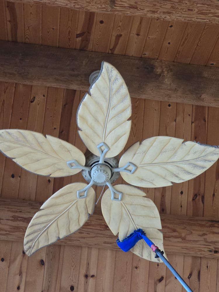 A person cleaning a leaf-shaped ceiling fan with a blue duster against a wooden ceiling backdrop.