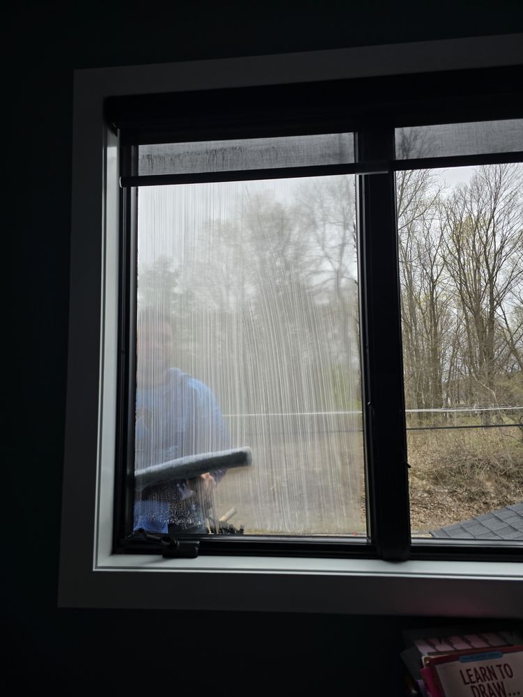 Window with a black frame, a roller shade at the top. A person is cleaning the glass.