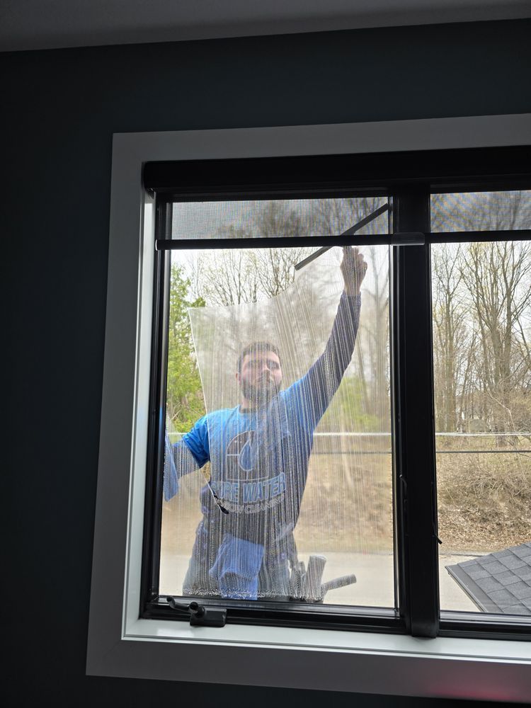 A man cleaning a window with a squeegee, inside a room with gray walls and a black-framed window.