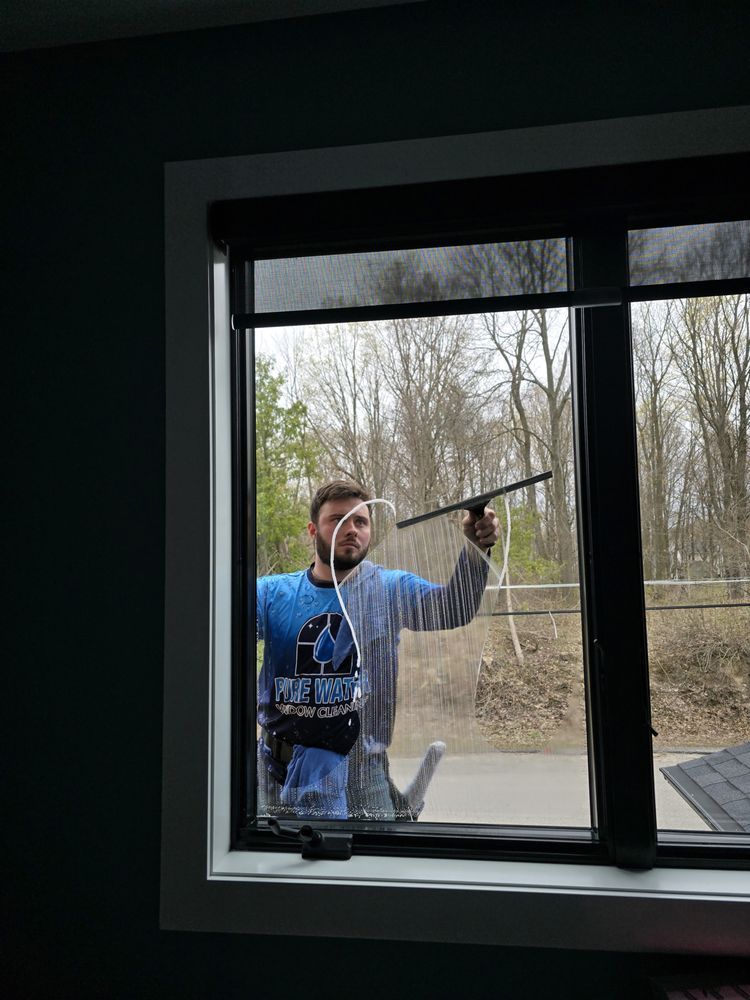 Two black-framed windows against a white wall, one being cleaned. The windows reflect a blue sky and green trees.