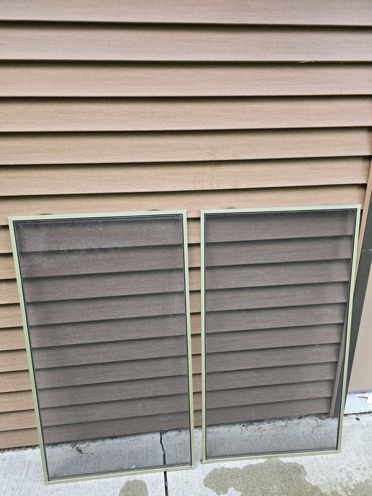 Two rectangular window screens resting against brown siding. The screens are dusty and damaged at the bottom.