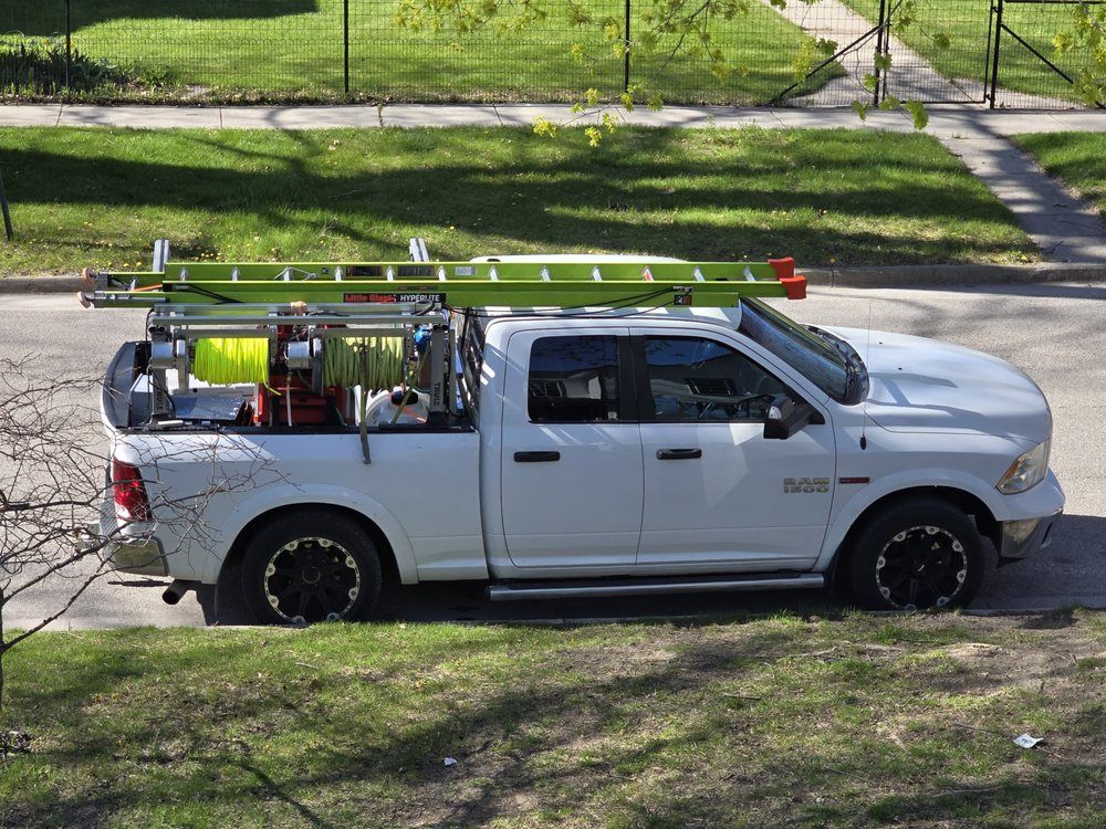 White pickup truck with ladder rack, parked on a street, carrying equipment; green ladder on top.