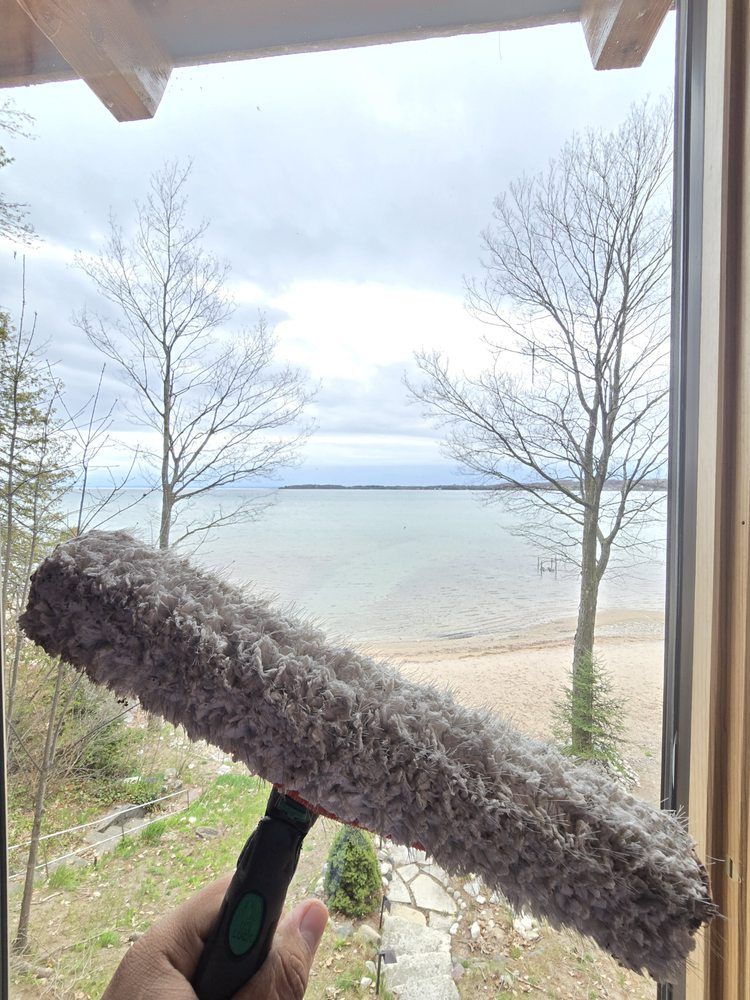 A hand holding a gray window cleaning applicator in front of a window with a lake and trees in the background.