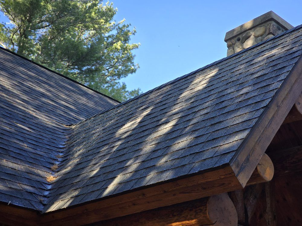 Close-up of a cabin roof with dark wood shingles and a stone chimney against a blue sky.