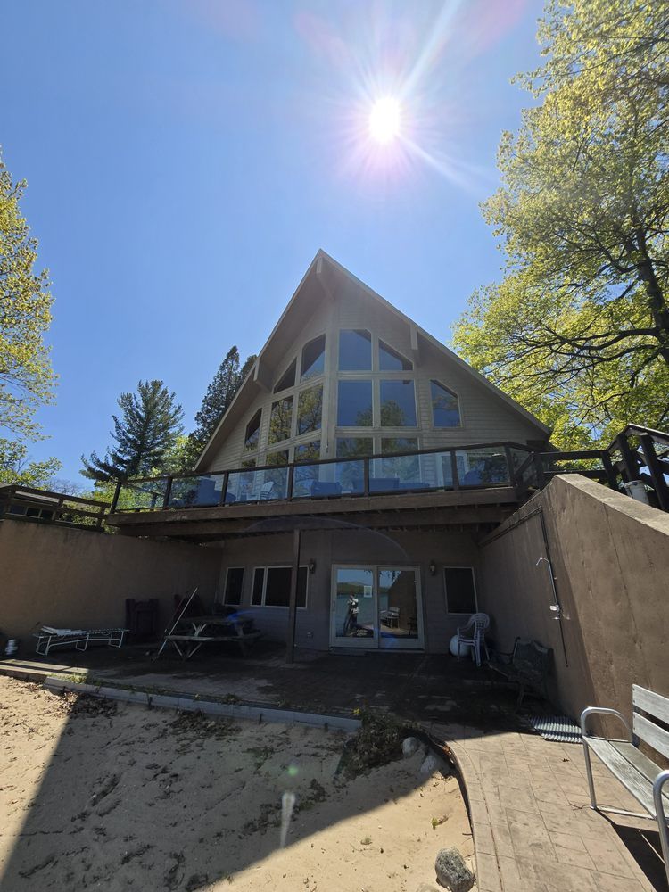 A-frame lake house with large windows and a glass-railed balcony, on a sandy beach under a bright blue sky.