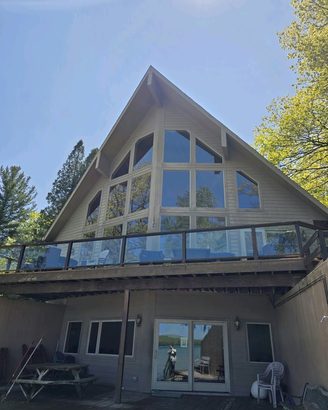 A-frame house with large windows and a deck overlooking a lake. The exterior is light-colored with a glass railing.