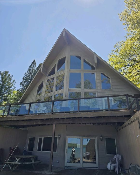 A-frame house with large windows and a deck overlooking a lake. The exterior is light-colored with a glass railing.