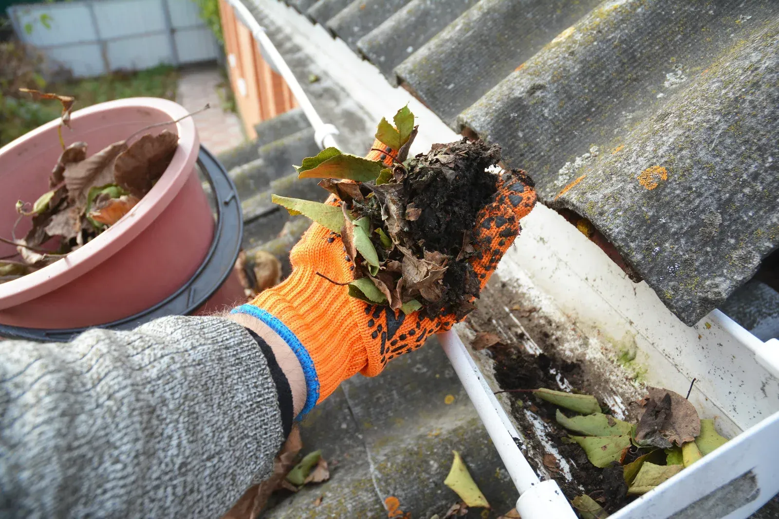 Person wearing an orange glove cleaning a gutter filled with leaves and debris on a rooftop.