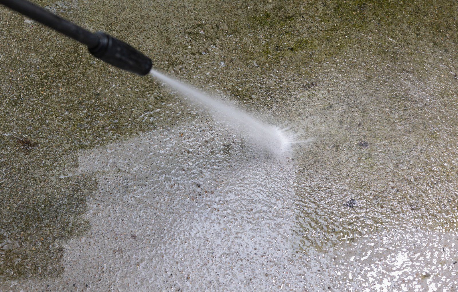 A power washer sprays water onto a concrete surface, cleaning it. The nozzle is black, and the concrete is wet.