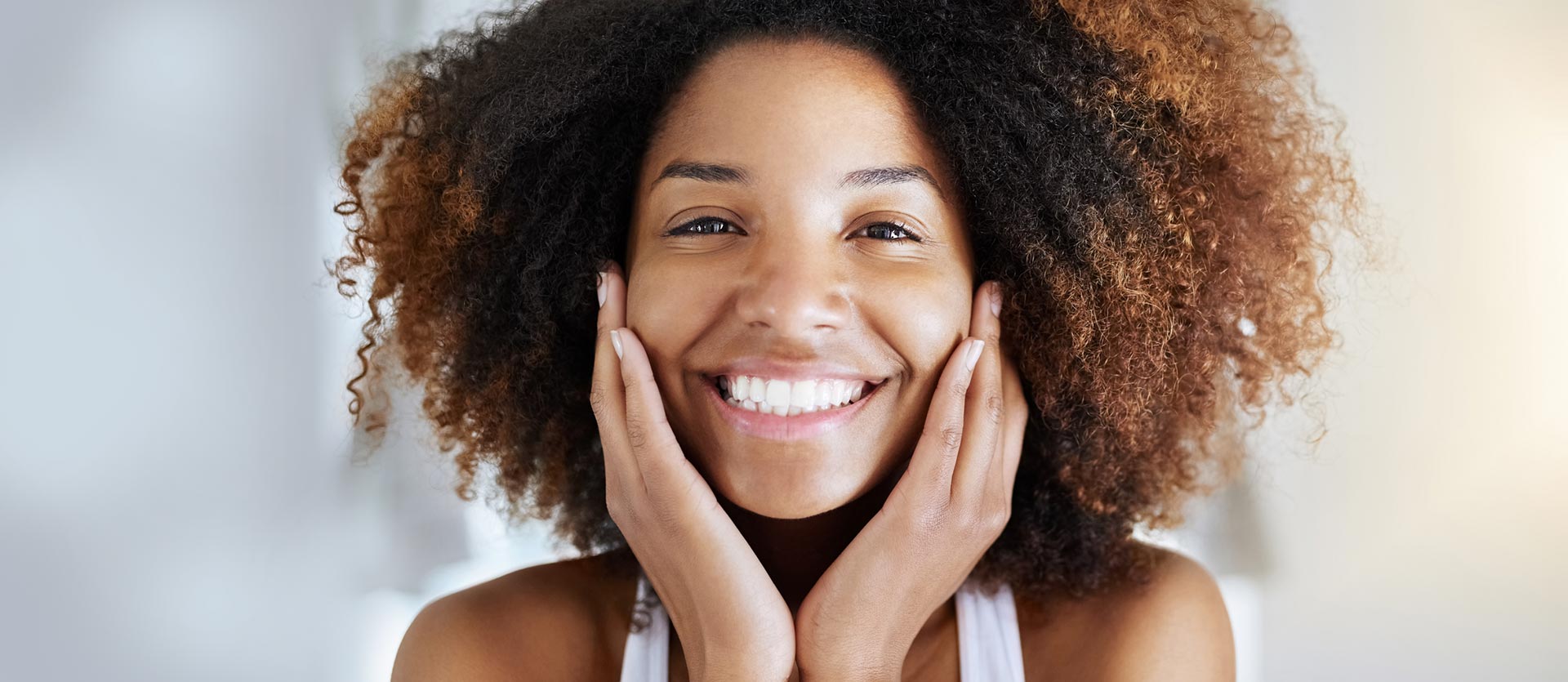 A woman with curly hair is smiling with her hands on her face.