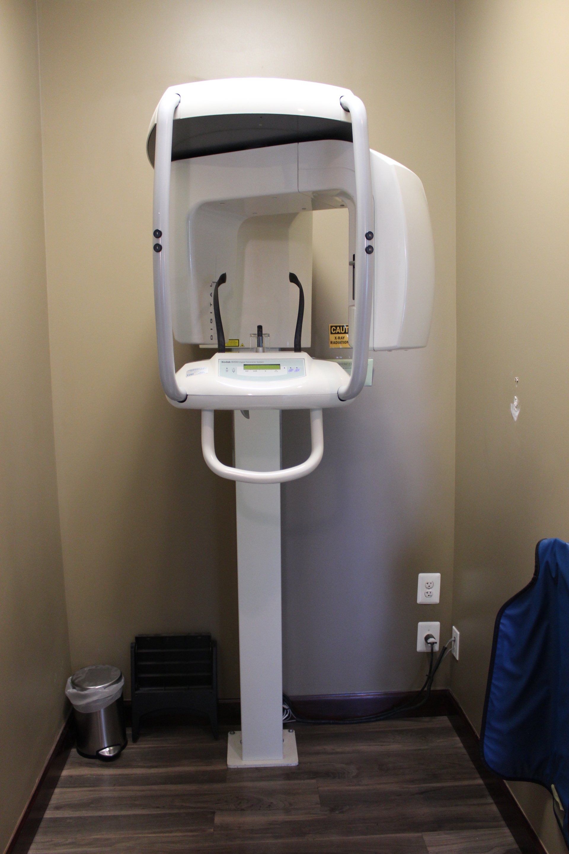 An x-ray machine is sitting in a small room next to a trash can.