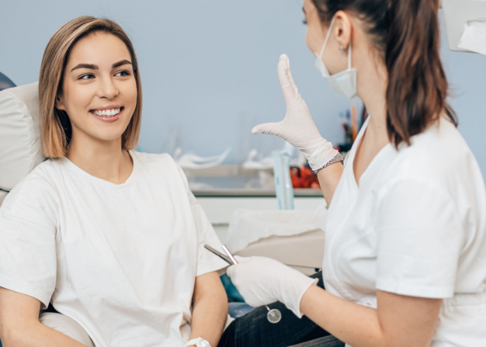 Woman smiling in a dentist's chair while dentist wearing gloves and a mask gestures.