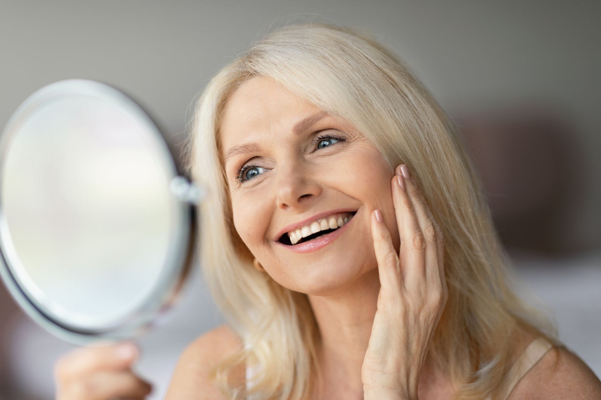 Woman with blonde hair smiling, looking at herself in a round mirror, touching her cheek.