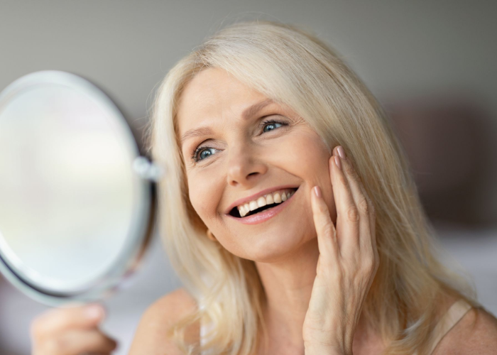 Blond woman smiling, looking in a mirror and touching her face. Indoors with soft lighting.