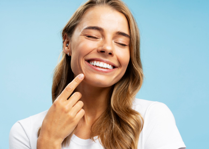Woman with eyes closed smiles, pointing at cheek with a finger. Light skin, wavy blonde hair, blue background.