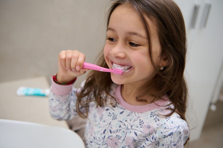 Girl brushing her teeth in a bathroom, wearing pajamas with pink toothbrush and toothpaste visible.