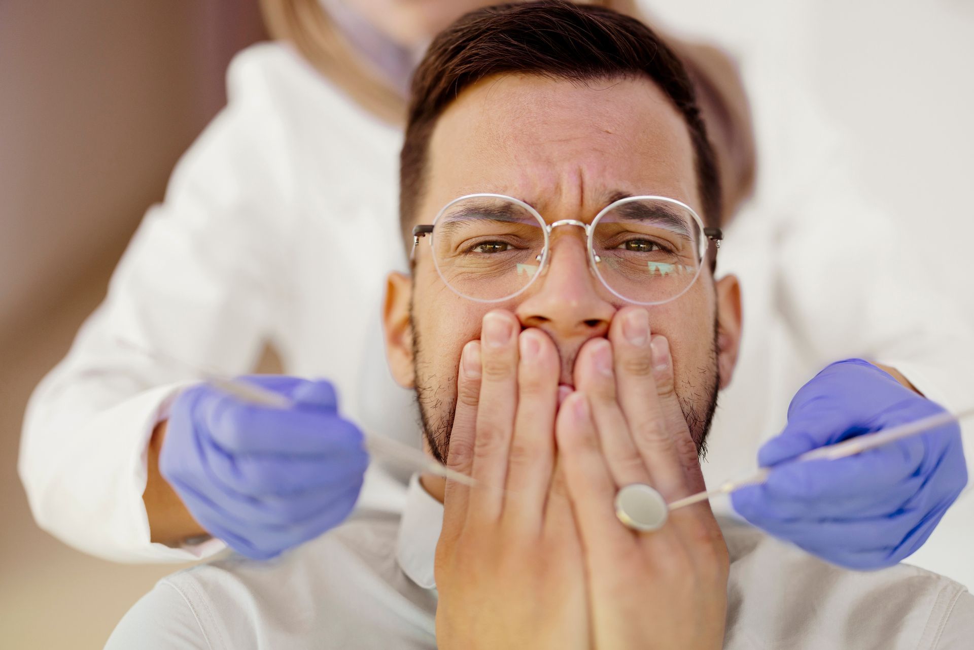 Man in glasses, frightened, covers mouth at dentist's office; dentist in gloves holds tools.