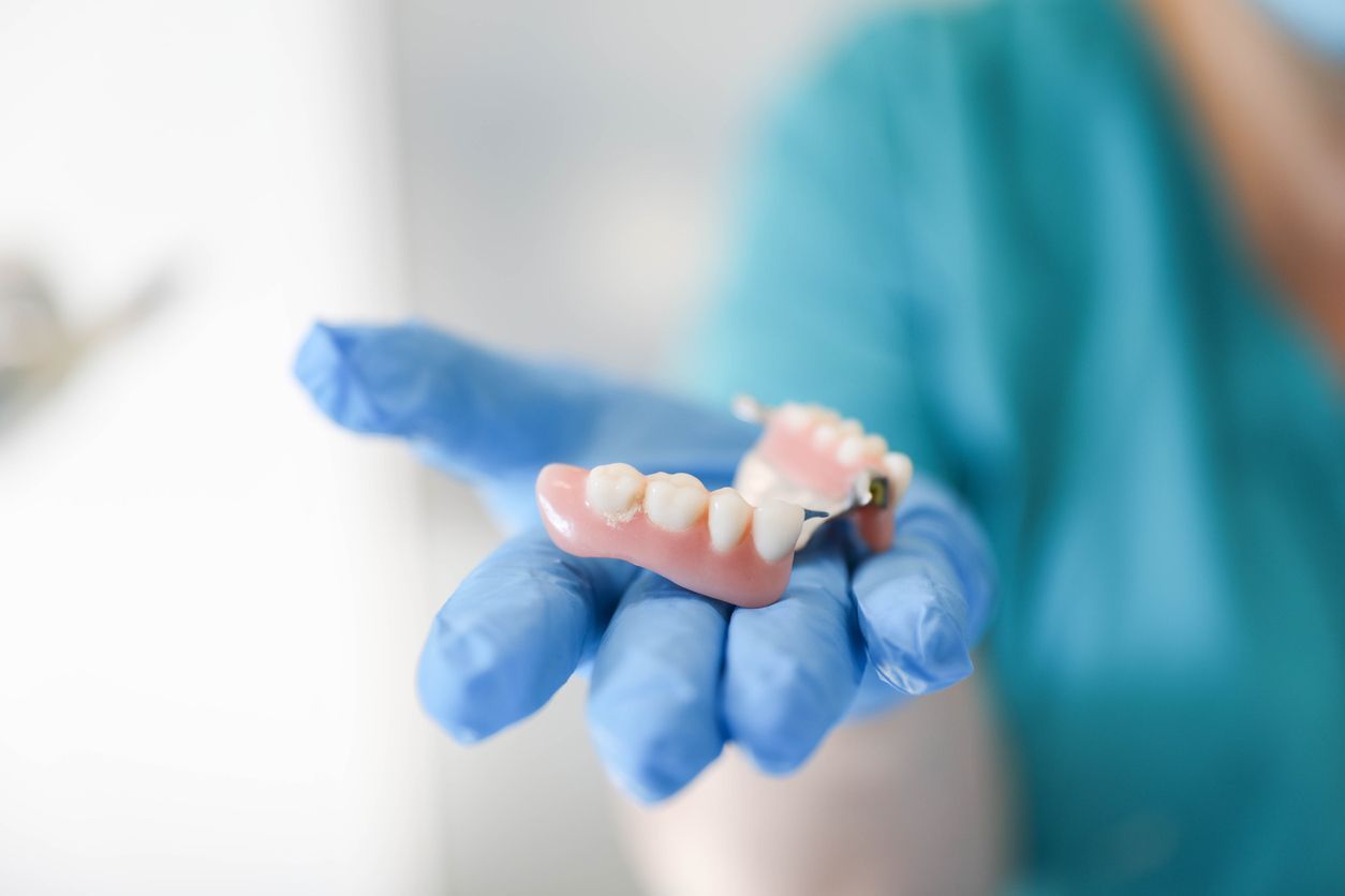 Dentist's gloved hand holding a partial denture, pink and white with metal clasps.