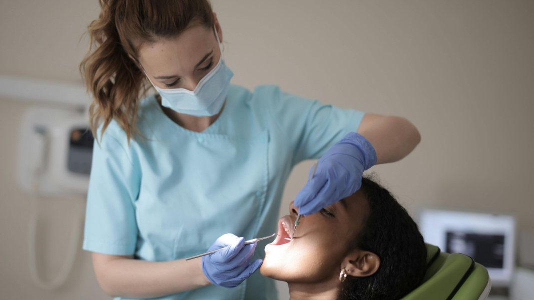 A masked dental professional in scrubs and blue gloves examines a patient's teeth with a metal tool in a clinical setting.