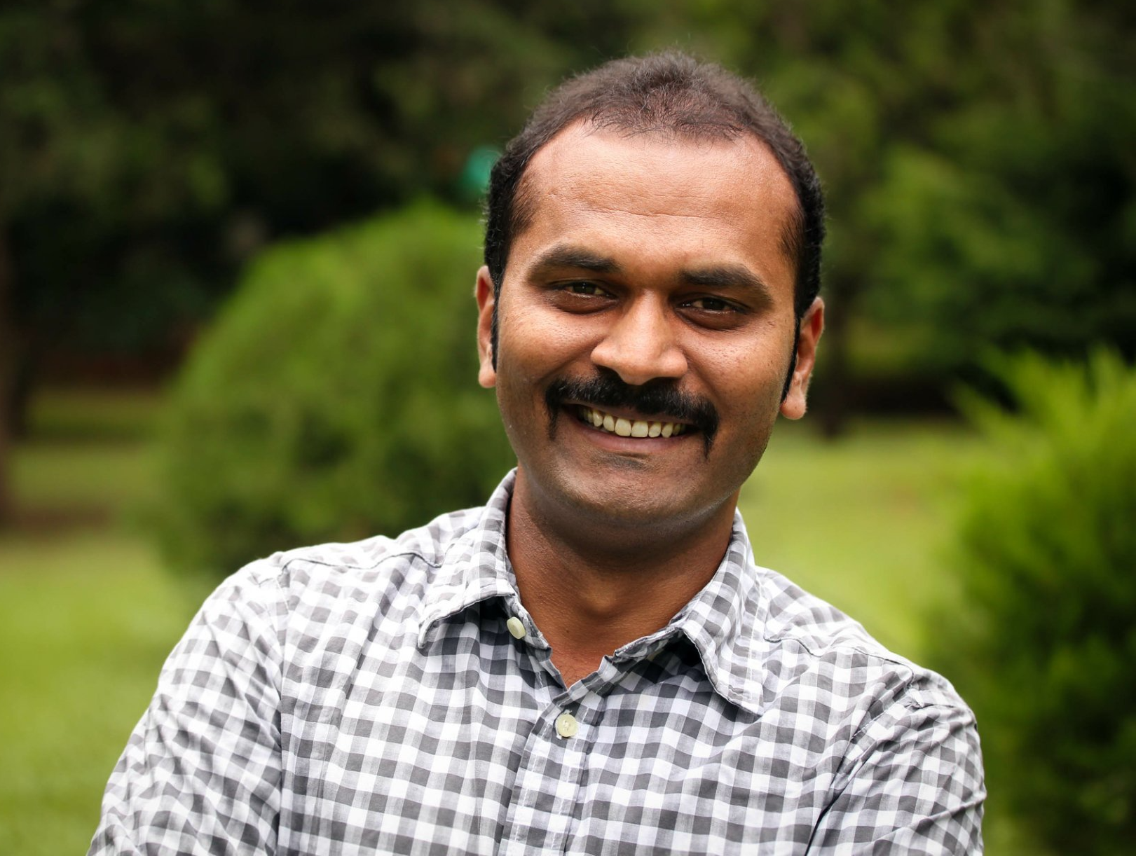 A man with a mustache is smiling and wearing a plaid shirt.