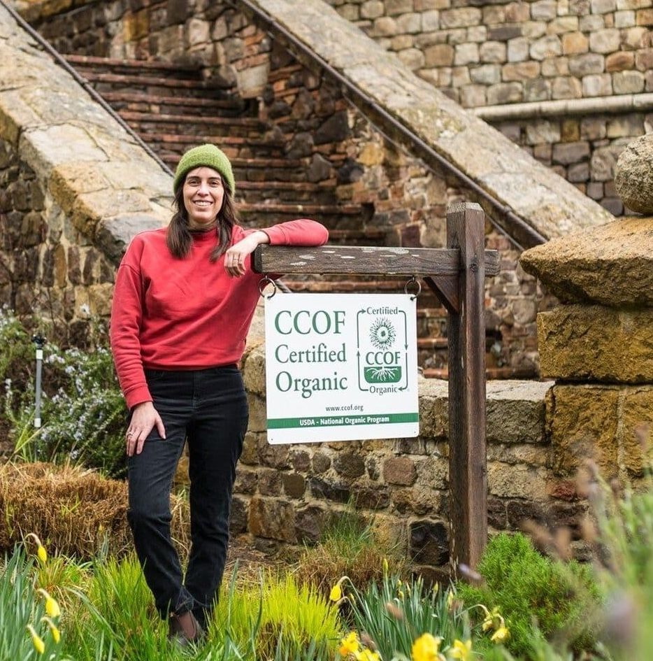 Woman in red shirt and green hat by CCOF Certified Organic sign, stone steps in background.