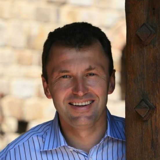 Man smiles, head and shoulders portrait; blue striped shirt, outdoors, wooden door frame, beige stone wall.