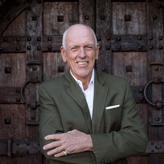 Man in green blazer smiles, arms crossed, in front of a weathered wooden door.