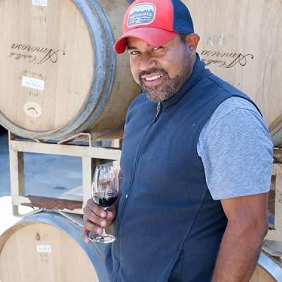 Man in vest and hat, holding wine glass near oak barrels.
