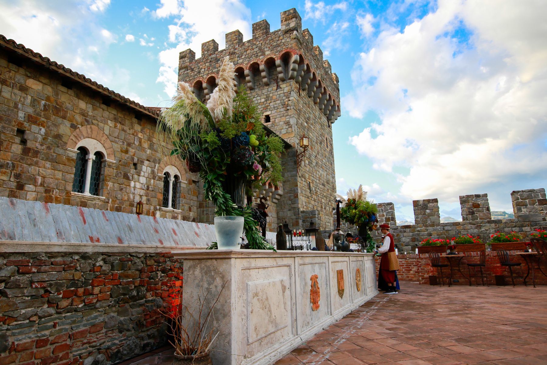 Outdoor dining area in a stone castle setting. Round tables with green cloths and chairs; barrel and mountain background.