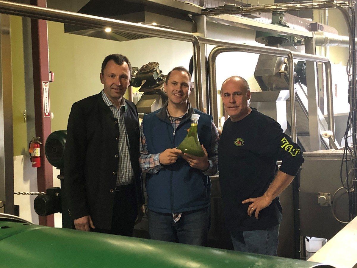 Three men inside a facility, one holding a bottle of olive oil.