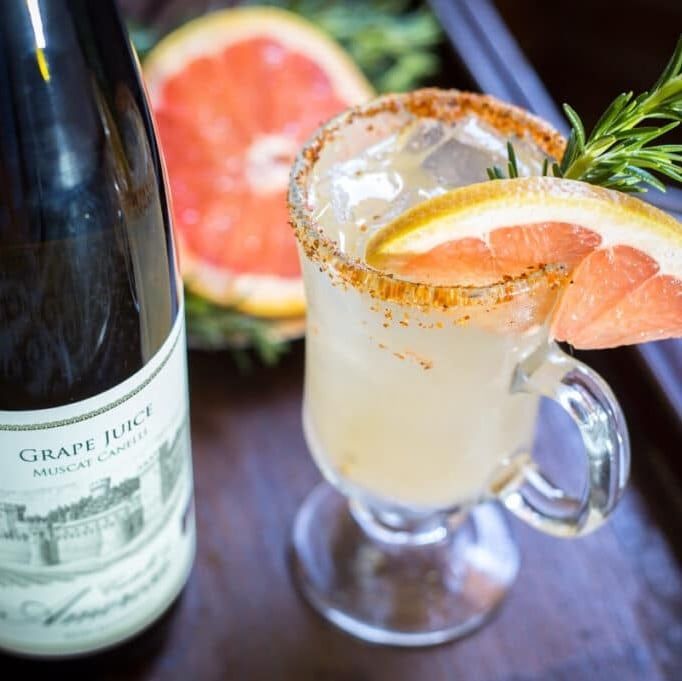 Grapefruit cocktail with rosemary sprig and spiced rim, served with grape juice bottle and grapefruit half.