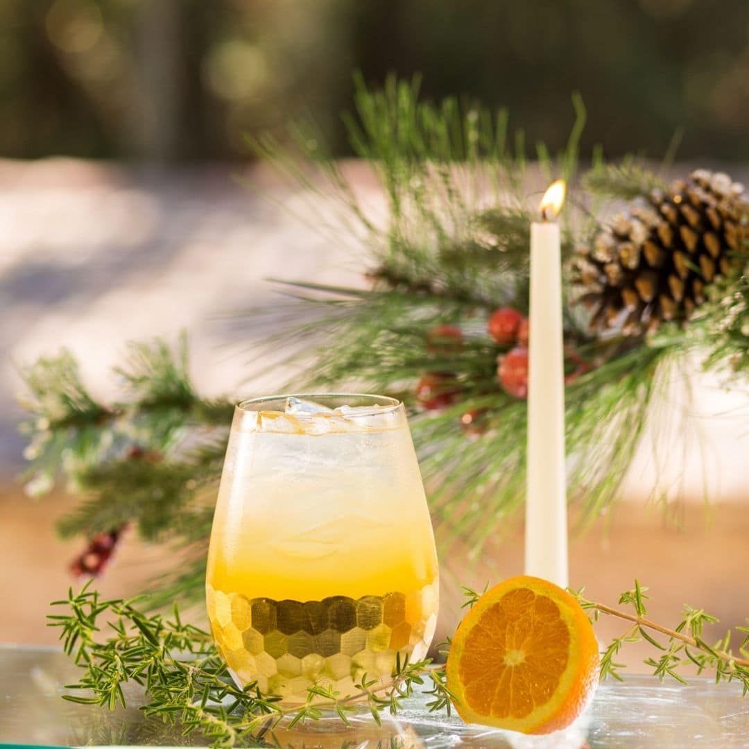 Glass of orange cocktail, halved orange, candle, pine boughs, and pinecone on a table.