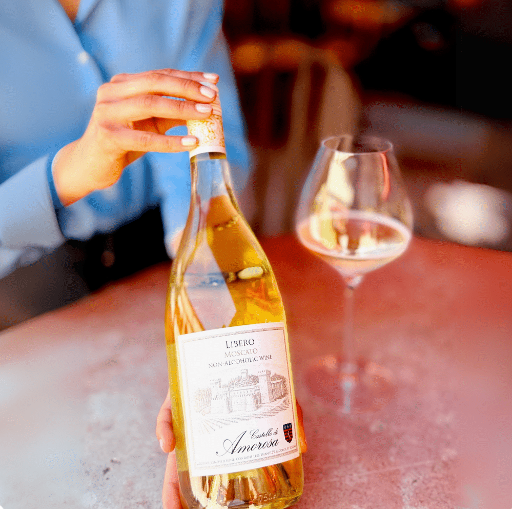 A hand holds an open bottle of white wine next to a filled wine glass on a red table.