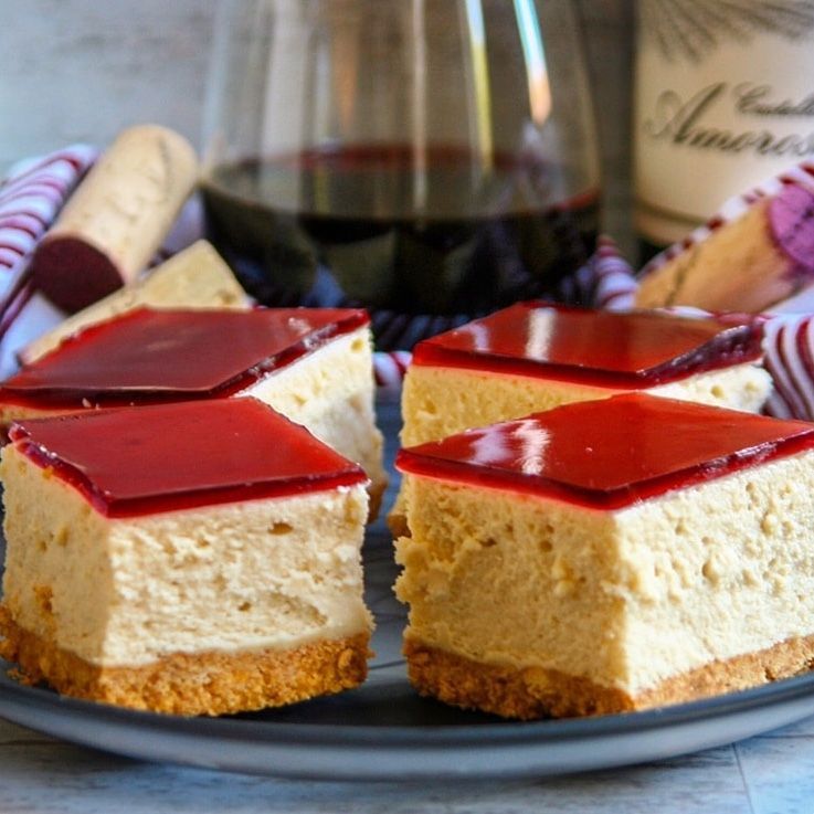 Cheesecake squares on a plate, topped with red glaze, in front of a wine bottle and glass.