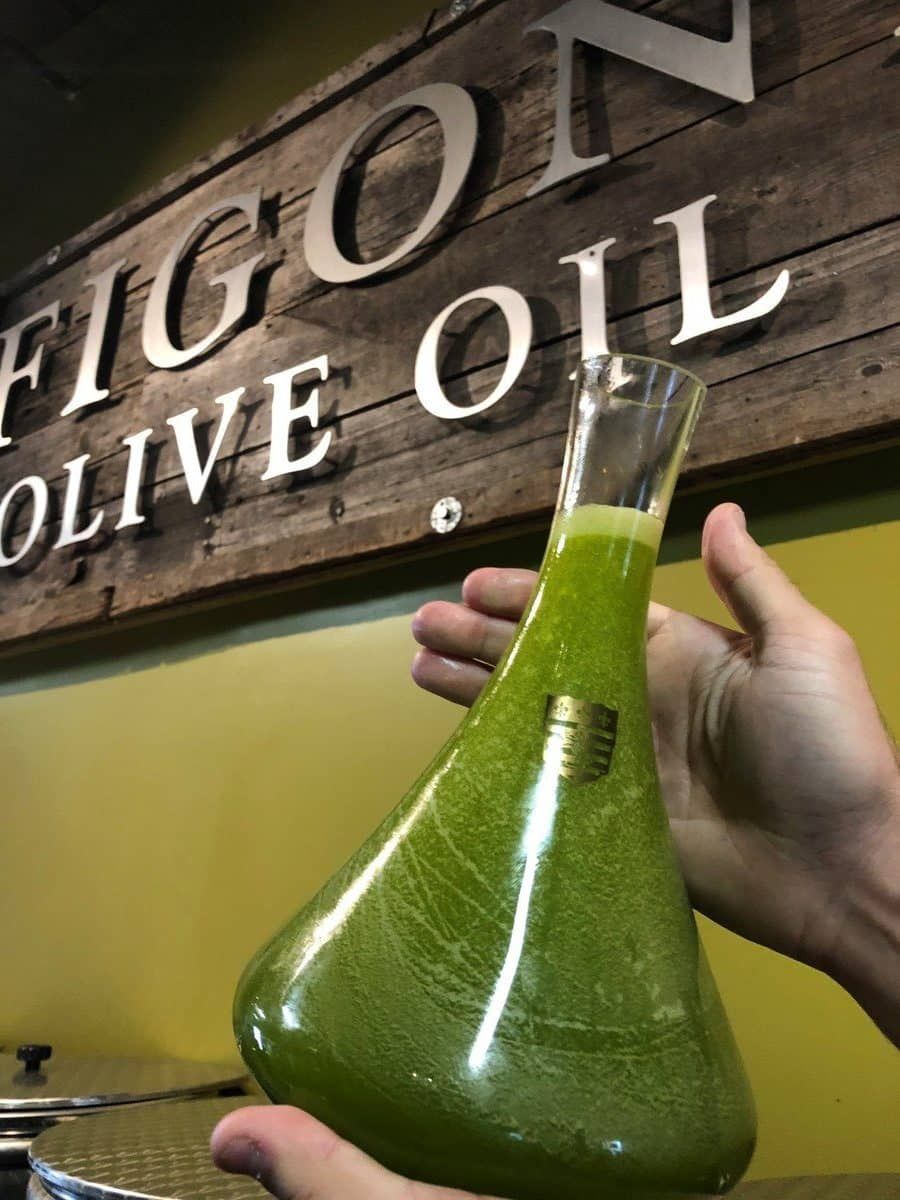 Person holding a glass container filled with olive oil with a sign in the background.
