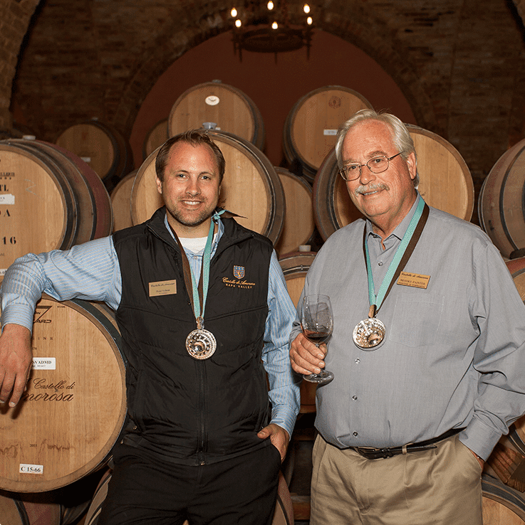 Two men in a wine cellar, wearing medals and standing near barrels. One holds a wine glass.