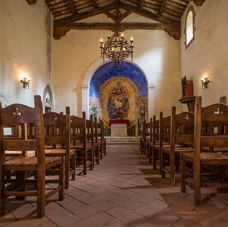 Interior of a chapel with rows of wooden chairs, altar with a painting, and a chandelier.