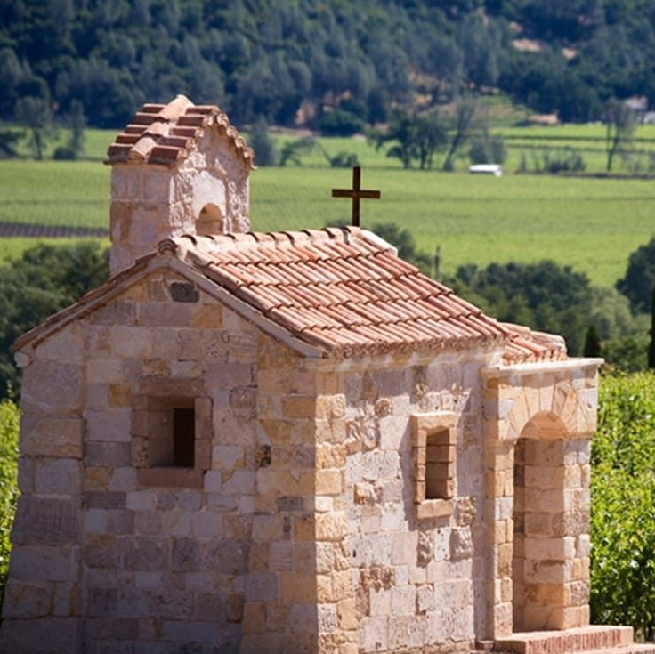 Stone chapel with cross on roof, set in a vineyard with a green backdrop.