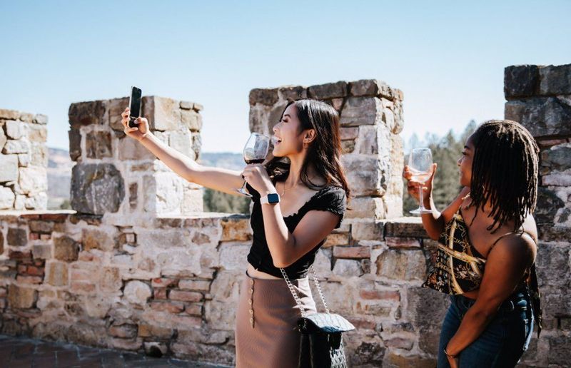 Two women taking a selfie on a stone wall, one holding a glass of red wine, outdoors in sunny weather.