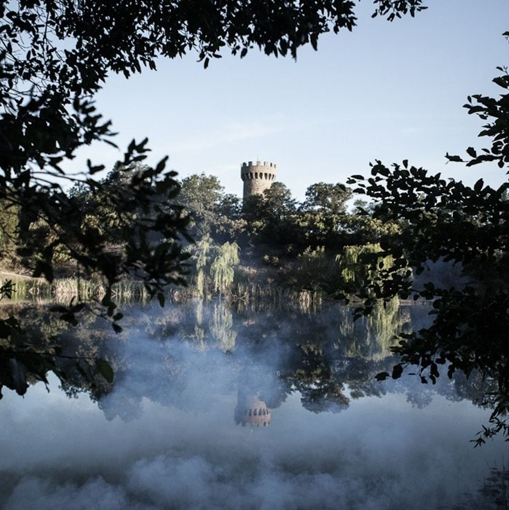 Stone tower reflected in misty lake, framed by tree branches.