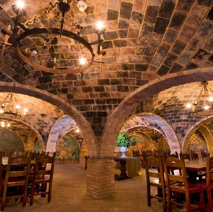 Brick-walled dining room with arched ceilings, tables, chairs, and chandeliers.