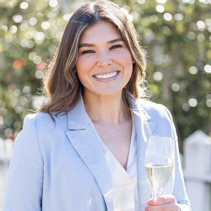Woman smiling, holding a champagne glass outdoors, wearing a light blue blazer.