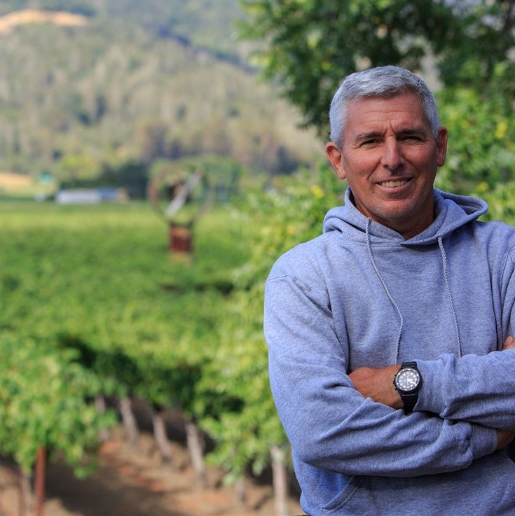 A person in a gray hoodie with arms crossed, standing in front of a vineyard with hills in the background.