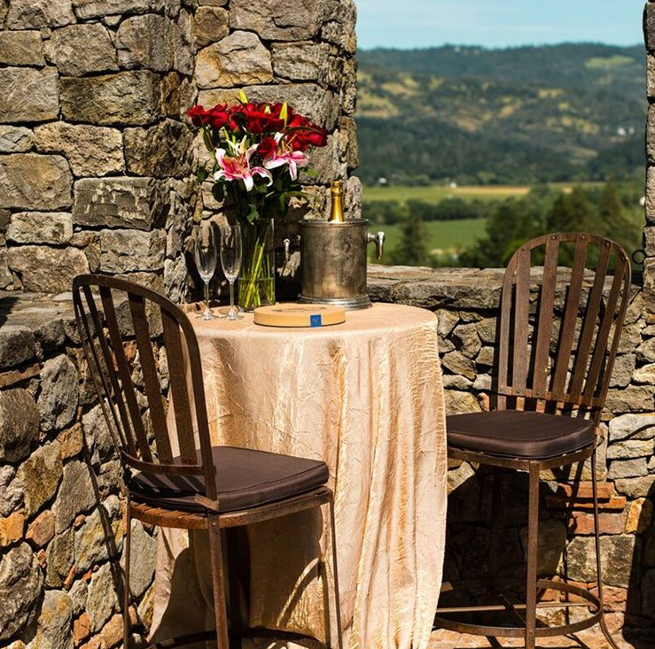 Outdoor dining set: table with gold cloth, two chairs, flowers, champagne, stone wall, scenic view.