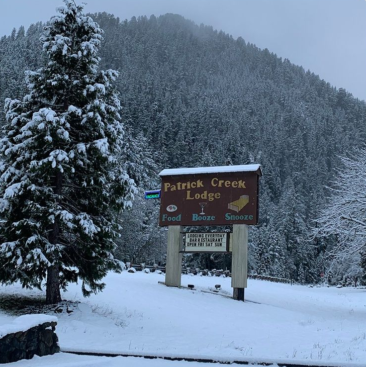 A wooden sign for Patrick Creek Lodge stands in a snowy, mountainous landscape next to a snow-covered evergreen tree.