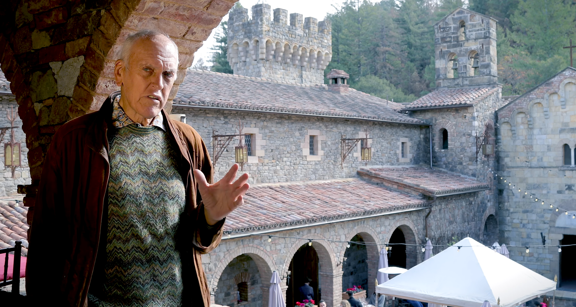 Man gesturing in front of a stone building with turrets and arched openings. Sunlight, outdoor setting.