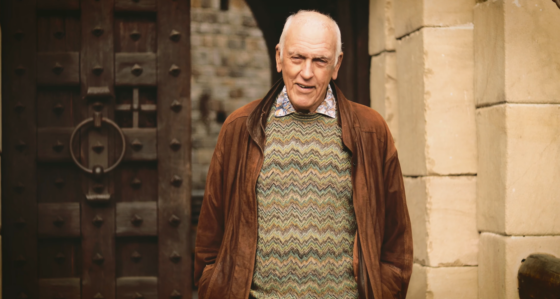 Man in brown jacket and sweater, stands in front of a large wooden door in a stone building.
